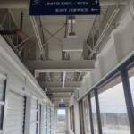 Long hallways with large windows on the right that let in a lot of natural light. To the left are various indoor storage units with white roll up doors. Hanging from the ceiling is a branded sign that directs customers to where their storage units are