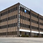 Wide angel photo of the 4 story storage facility with a large sign of the Defender Storage logo. THe building is rectangular, brown, and made of a mix of brick and stone